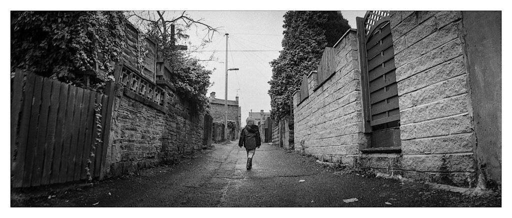 Alleyway at back of terraced houses. Small child walking down the middle of the panoramic frame