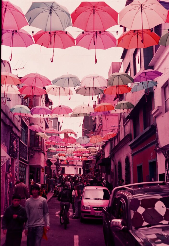 A narrow street in Bogotá decorated with umbrellas overhead. The film has a light purple to pink cast.