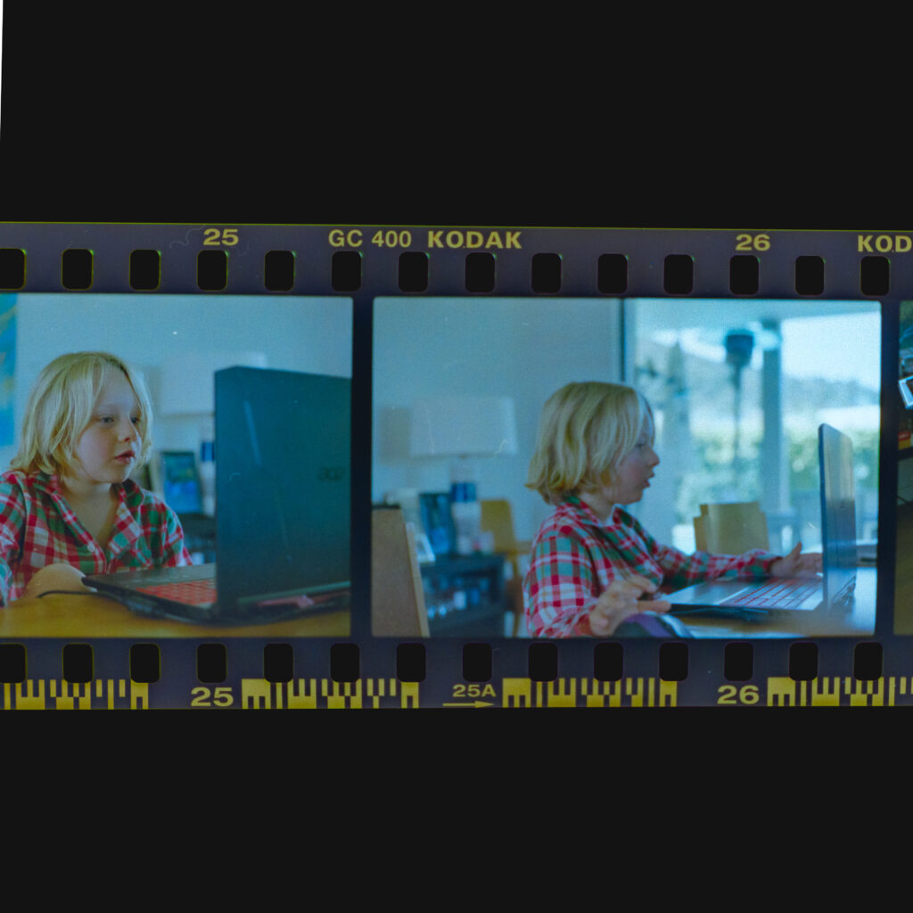 Close up scan of a 35mm film strip taken with a DSLR camera. Two frames are visible, both showing a blond boy playing on a laptop at a wooden table.