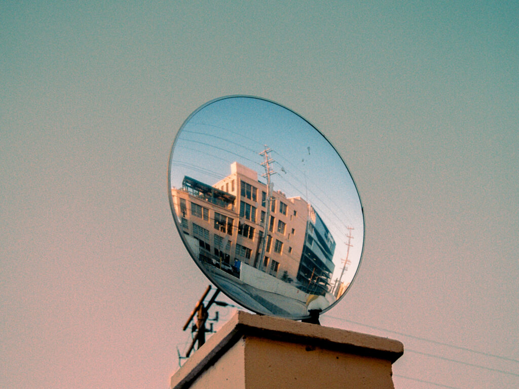 Blocky building in pinkish, dusky light captured in a circular mirror outside a parking lot. Most of the frame is sky, with the mirror at the center.
