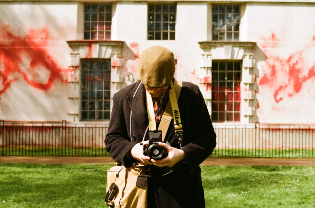 Jimmy taking a picture on a medium format camera, with a building behind with red paint on.