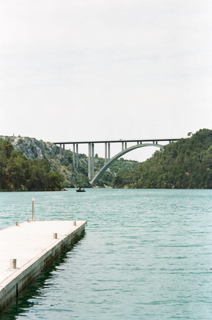 A small pier jutting out to a large body of water. In the distance, a large metal bridge and green hills either side. 