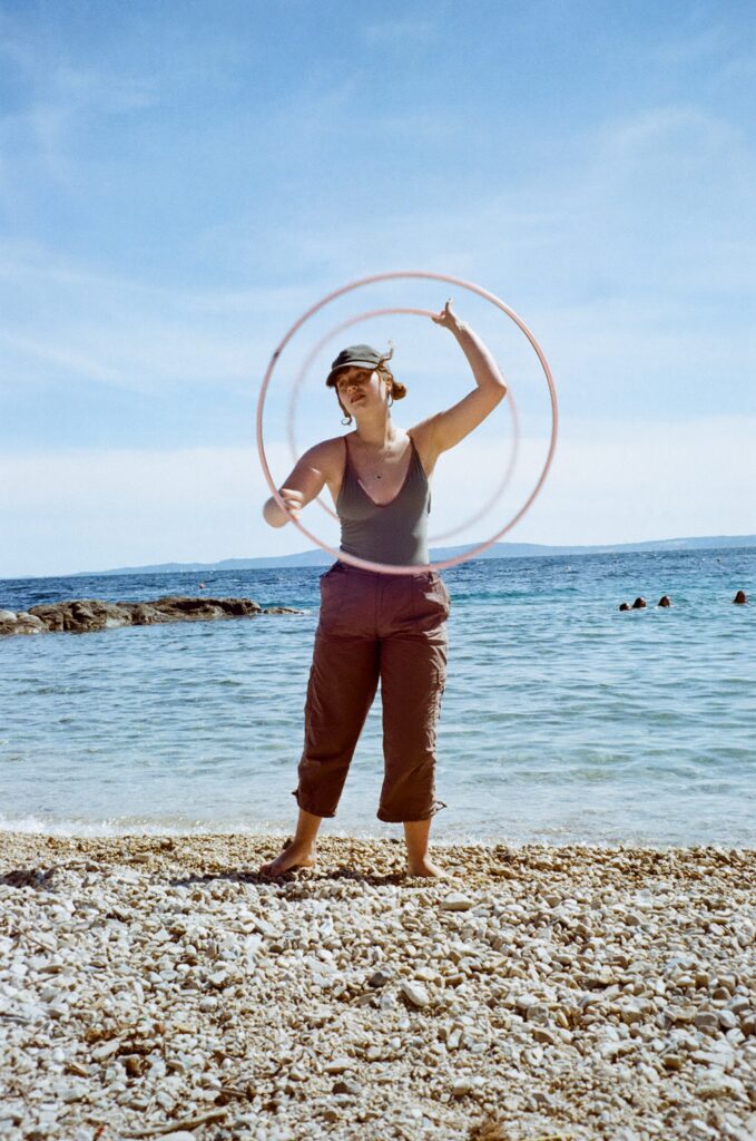 A young woman standing on a beach, one hula hoop spinning around her face and one spinning behind her back.