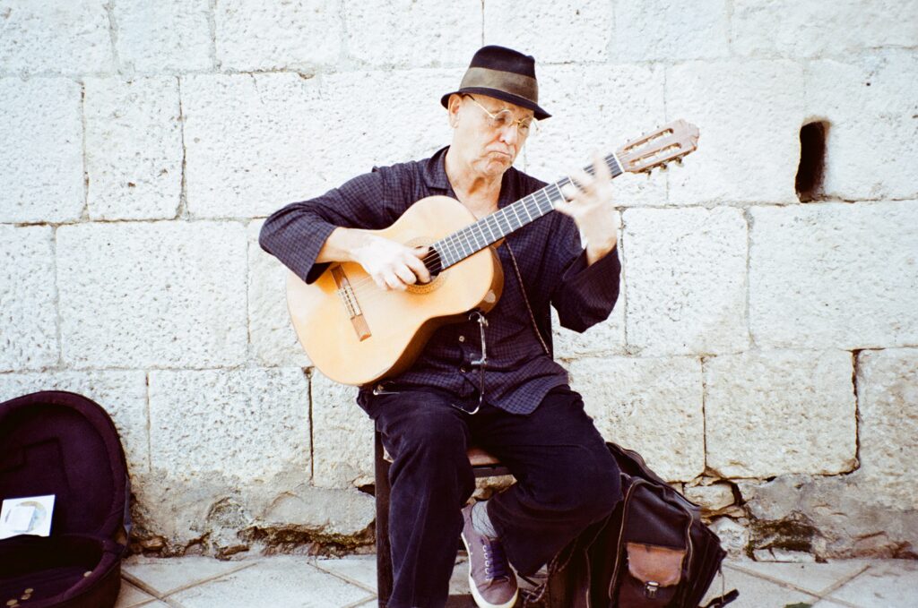 An old man playing the guitar in front of a light stone wall. 