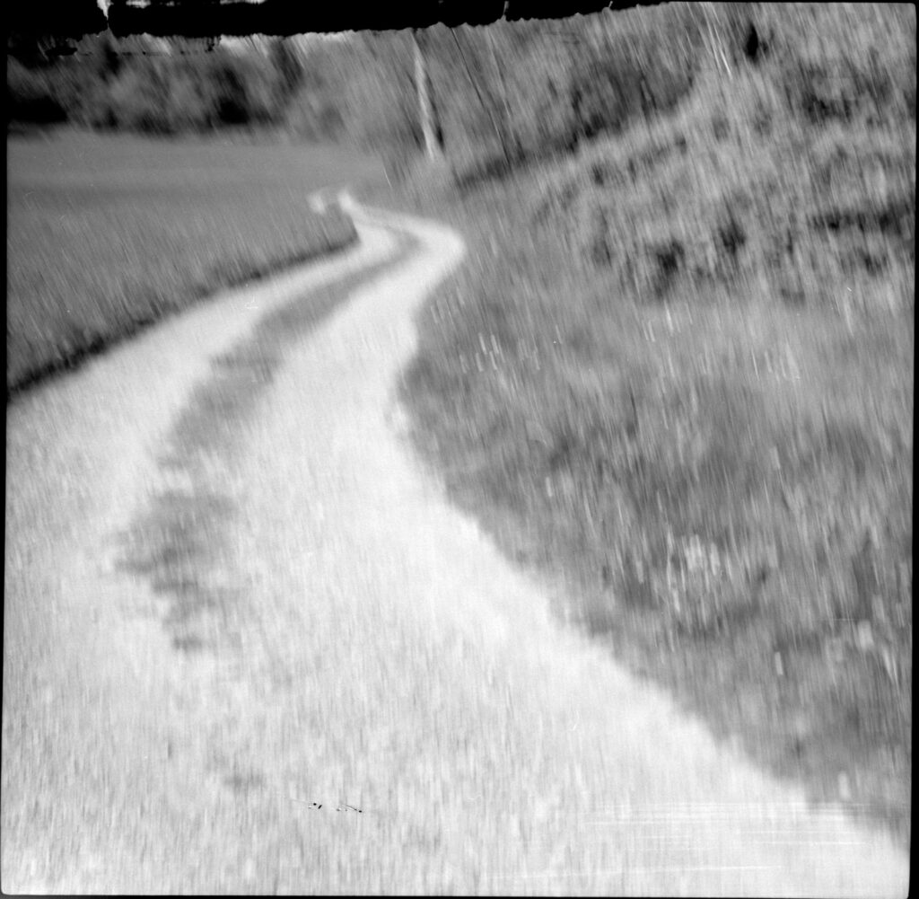 a black and white photo of a dirt road. the photo is blurry from motion as the photographer moved to fix the camera, and there is a black, splotchy area to the top of the frame where the film didn't develop properly