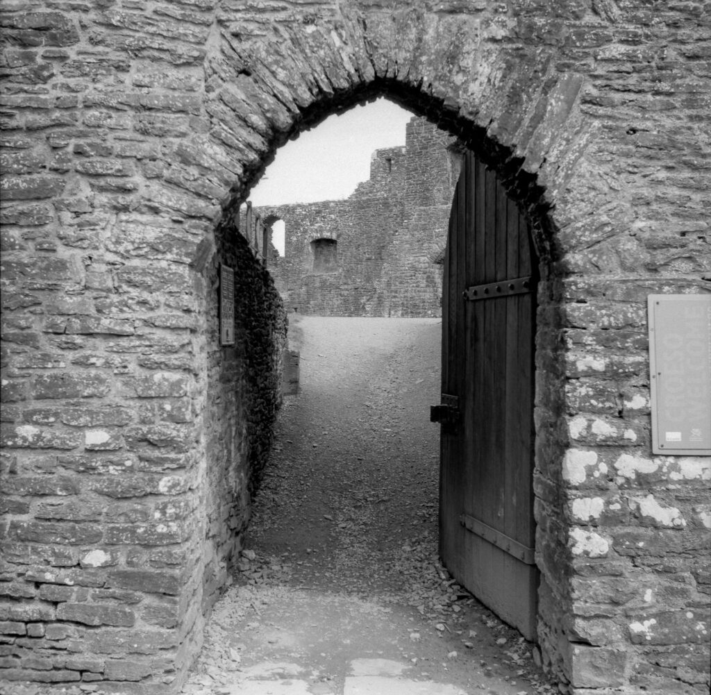 A black and white photo of a stone arched doorway with a large wooden door, leading into a ruined castle courtyard