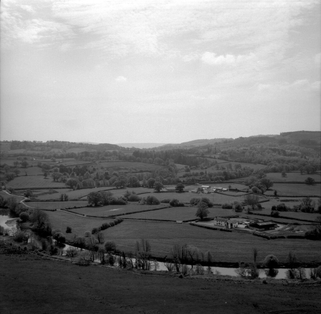 a black and white photo of a landscape, a river running along the bottom of the frame and farm buildings are visible