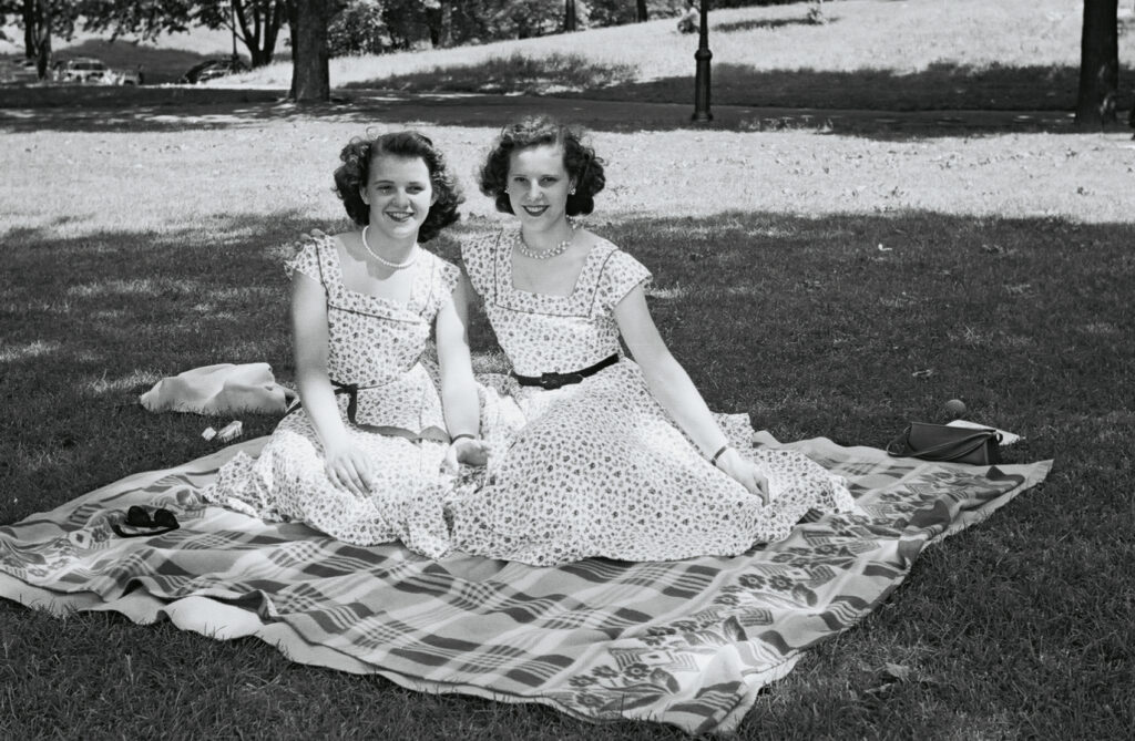 A black and white photo of 2 young women in dresses sitting on a blanket on the grass. 