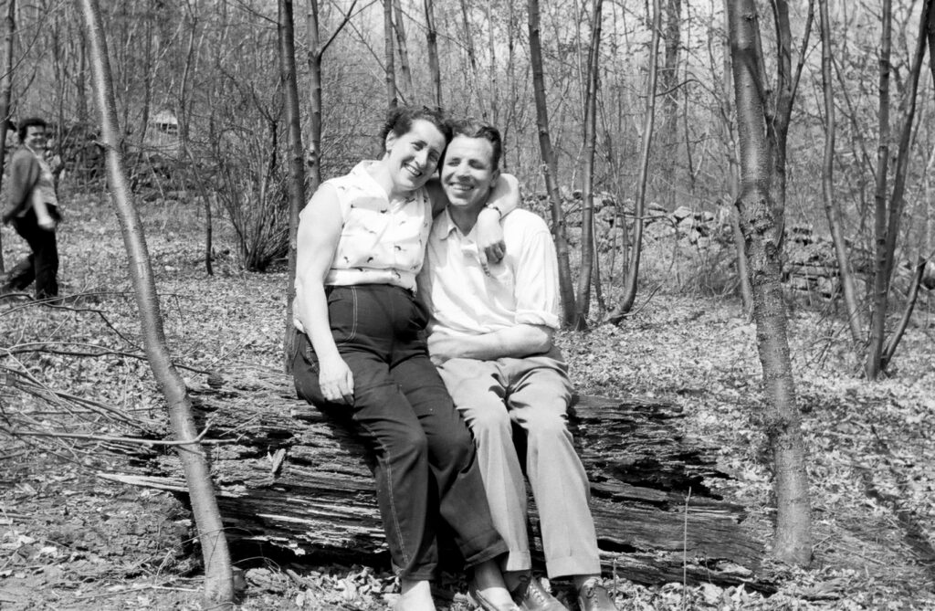 A black and white photo of a man and woman smiling and sitting next to each other on a log. 