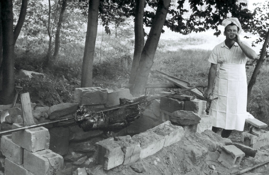 A black-and-white photo of a man cooking a roast pig on a spit over an open fire