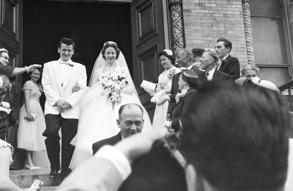 A black and white photo of a bride and groom exiting a church with people on the stairs looking at them.