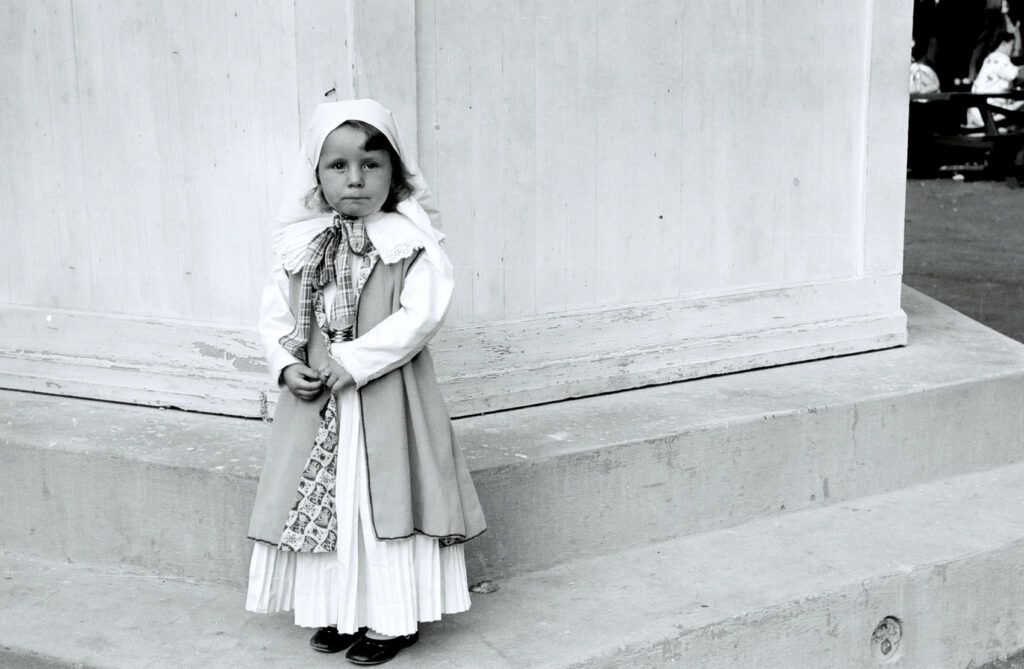 A black and white photo of a young girl in traditional Gottscheer dress standing by herself. 