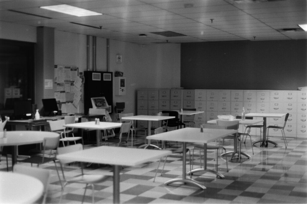 Black and White image of a break room with a variety of small tables, a checkered floor, and a long row of filing cabinets along the back wall