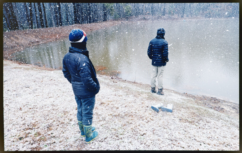 2 boys in winter jackets standing by a pond in the snow.