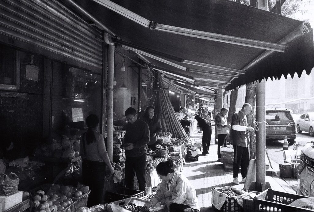 A row of shopfronts opening onto a covered pavement. There are shop staff and shoppers.