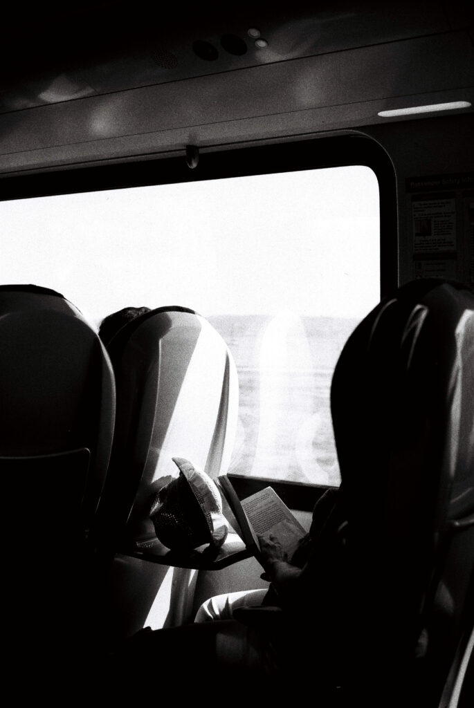 Black and white photo of a person reading on a train. The book and a hat are visible on the tray table.