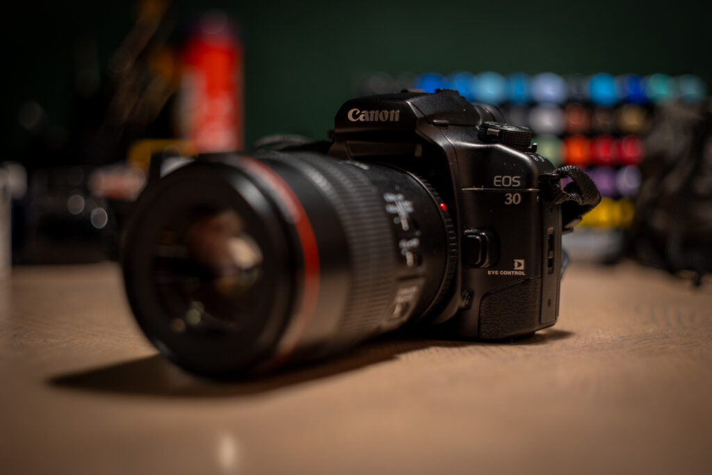 an image of a camera with a lens sitting on a desk. the lens has a red ring, and is quite large. the visible text on the camera says "Canon EOS 30". behind the camera is a colourful blur of pens