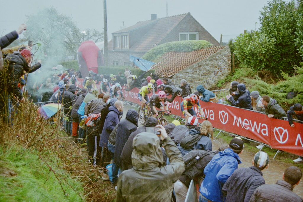 Green smoke rises from the left of the photo while cyclists ride along a cobbled street running through the image. Fans wearing raincoats behind barriers take photos and cheer riders on.