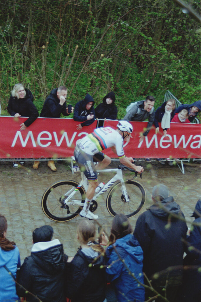 A lone cyclist in a white jersey with multi-coloured stripes rides along a cobbled street in the cnetre of the image, while fans behind red barriers scream and take photos. 