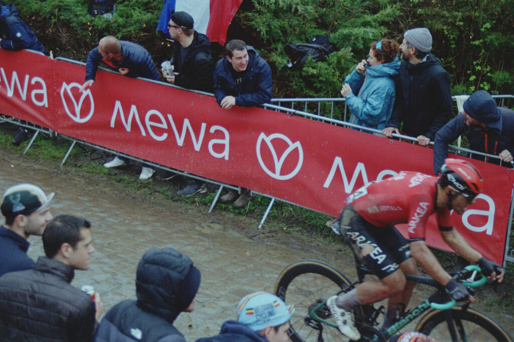 In top centre, a supporter cheers on a rider who is disappearing into the bottom right corner of the image. A French flag is draped behind the supporter and his friends, who are looking the other way. 