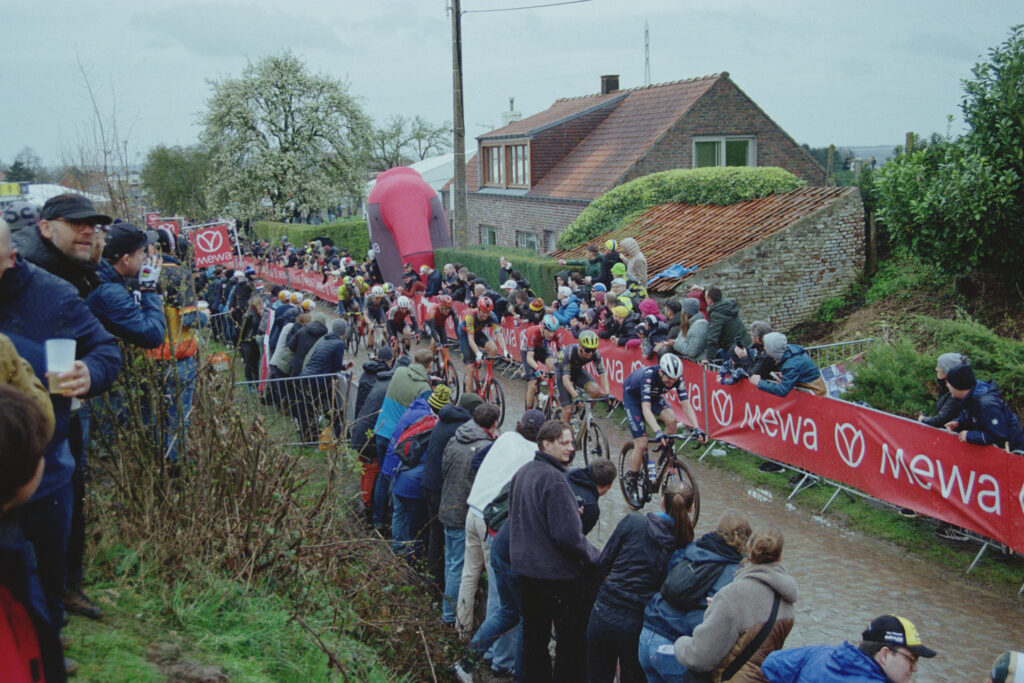 Cyclists ride along a cobbled road in single file while fans line barriers to cheer them on. In the background is a farmhouse and large inflated red cycling jersey. 