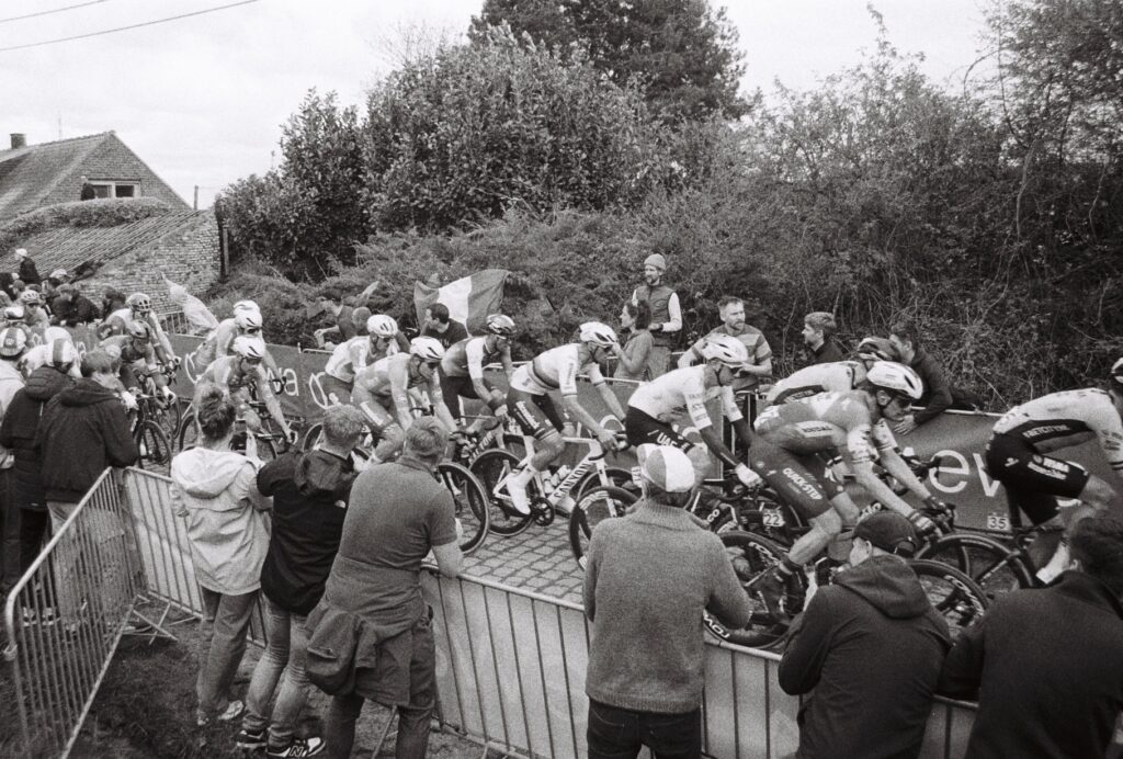 Racing cyclists ride at speed along a cobbled street while fans cheer on from behind barriers. At the centre of the photo a cyclist stands out in a white jersey with bands around the centre. 