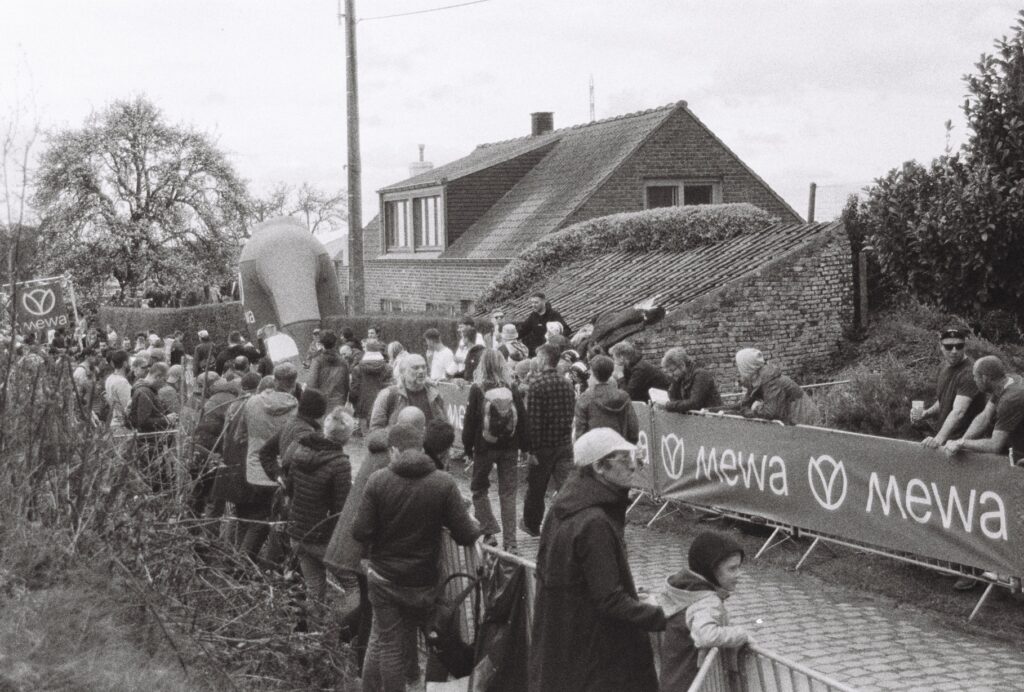 Two groups of fans on opposing sides of a cobbled street shout at each other over the top of a group of fans in walking along the street. The distinction between groups is not clear. 