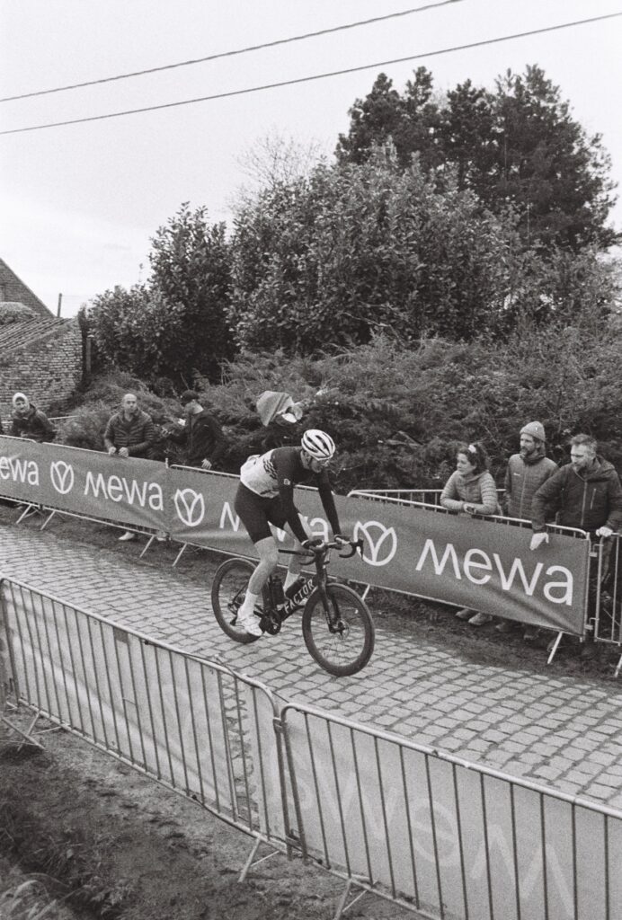 A lone cyclist rides along the cobbled street, while a couple of fans behind barriers cheer him on