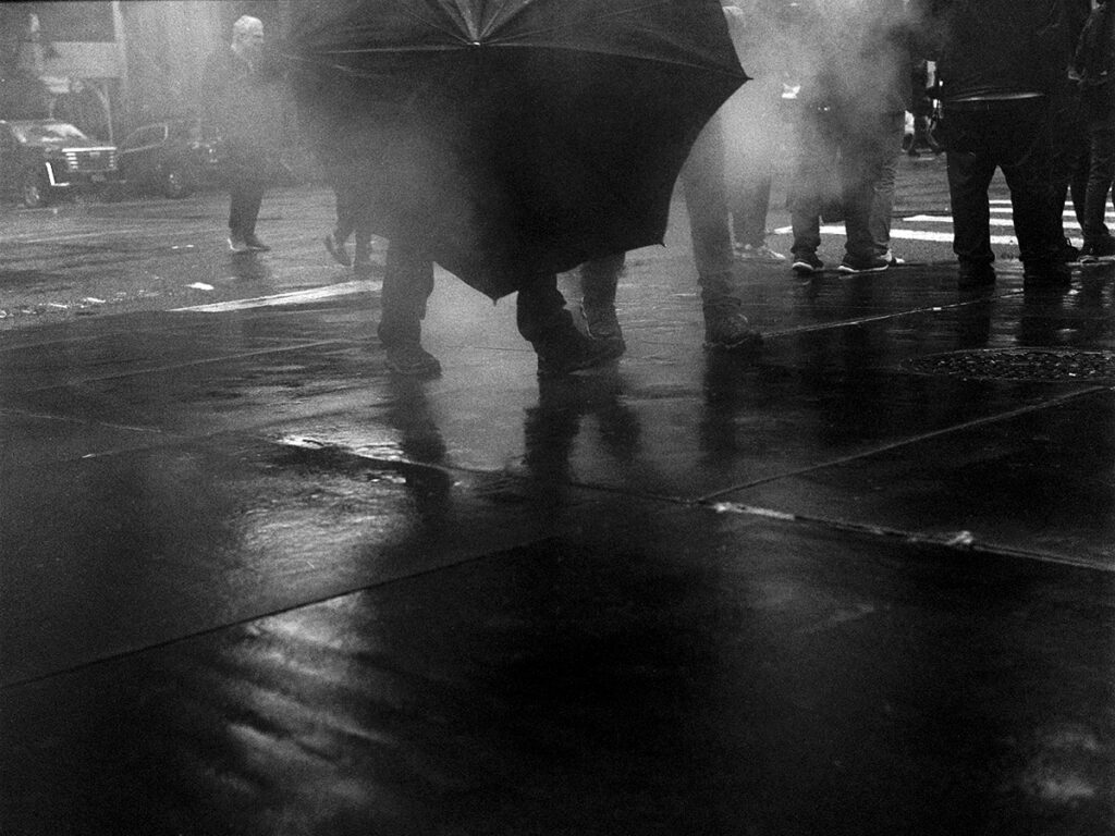 Black and white photo of a wet, shadowy street corner covered with mist and fog and intersected with a black umbrella and the lower legs of pedestrians.