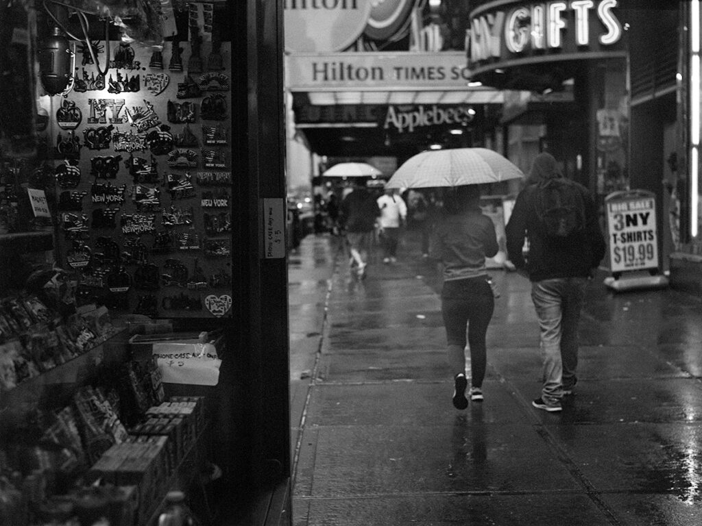 Black and white image showing partial interior of a New York City news stand on the left and a rainy sidewalk traversed by umbrella carrying pedestrians on the right.