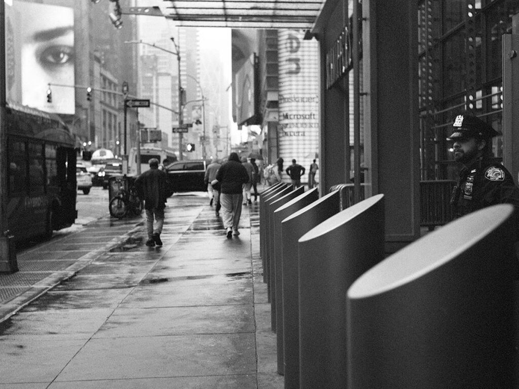 Wet sidewalks in front of the barricades beside the New York Times building in NYC, showing a police officer guarding the entrance to the building and a billboard with a face that is "watching" the officer from the left side of the frame.