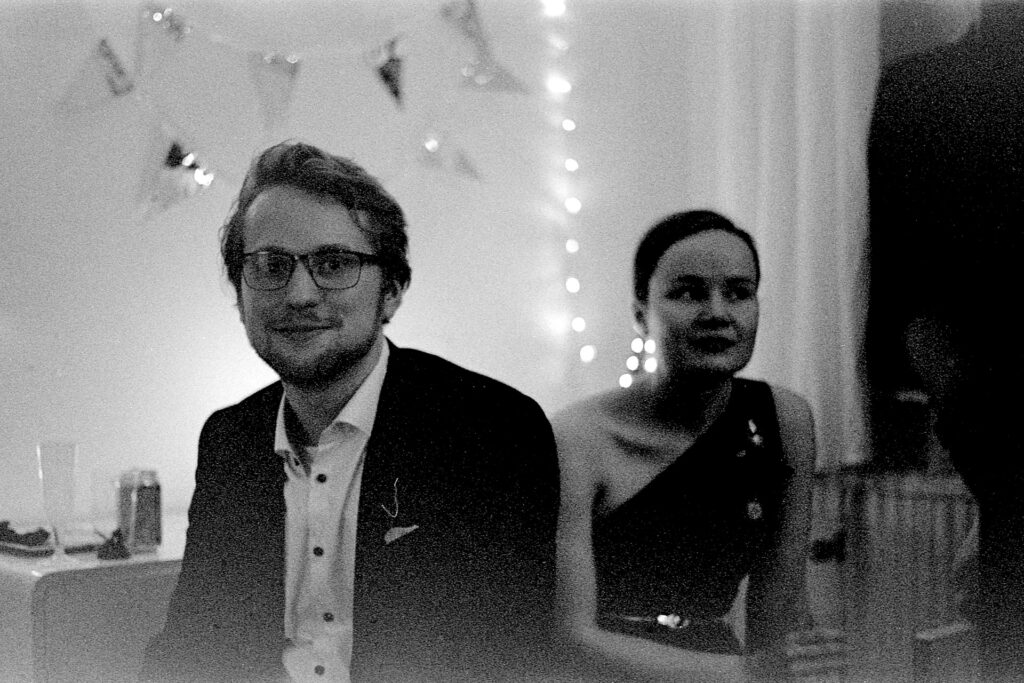 A black and white photograph of a young man and woman at a house party. The man's dressed in smart casual attire, and the woman is wearing an evening dress.