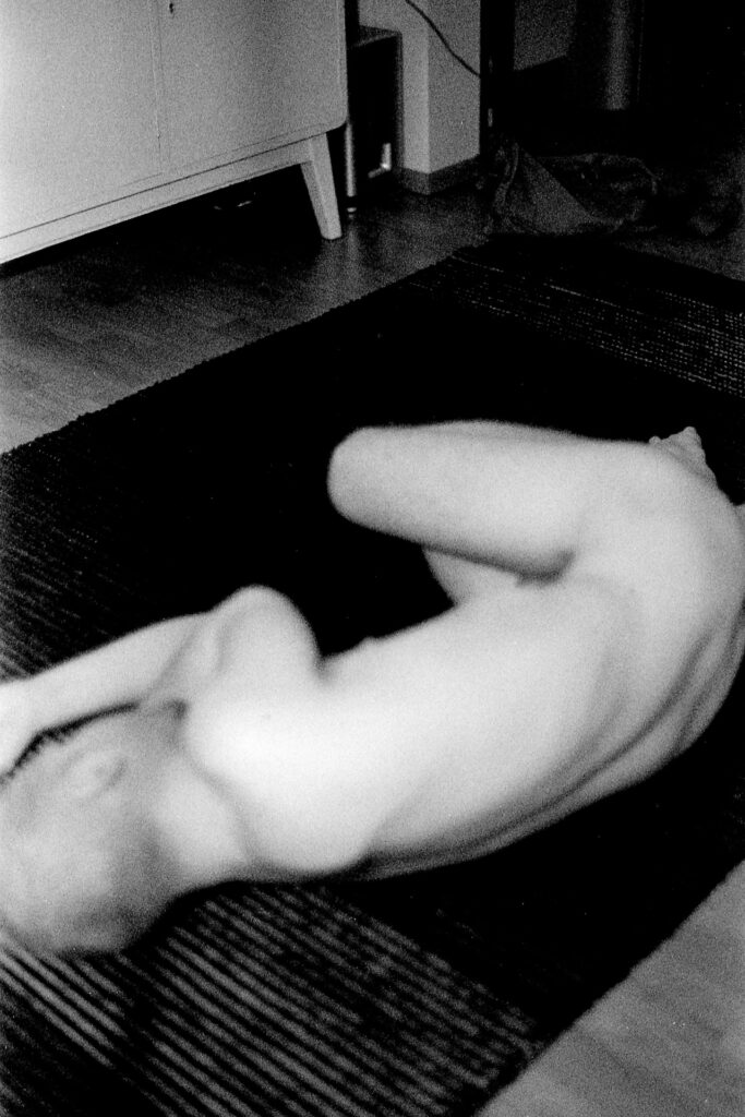 A black and white photograph of a naked man lying almost in a fetal position on a dark carpet. The man's back is towards the camera. The man is out of focus. 