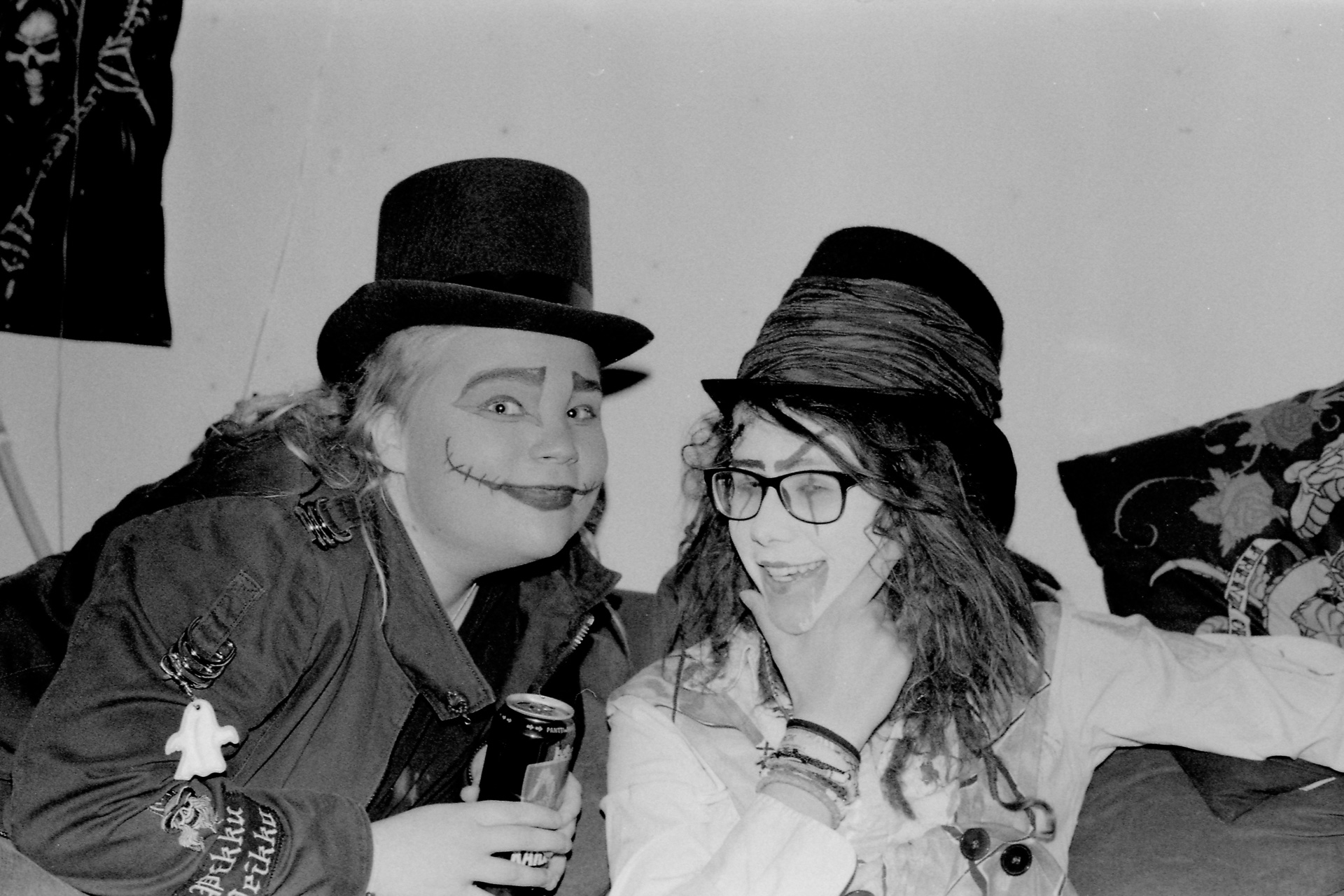 A black and white photograph of two women in Halloween costumes posing for the camera. First one's wearing a black top hat and face paint depicting scars around her mouth, the other is wearing a Mad Hatter costume of sorts.