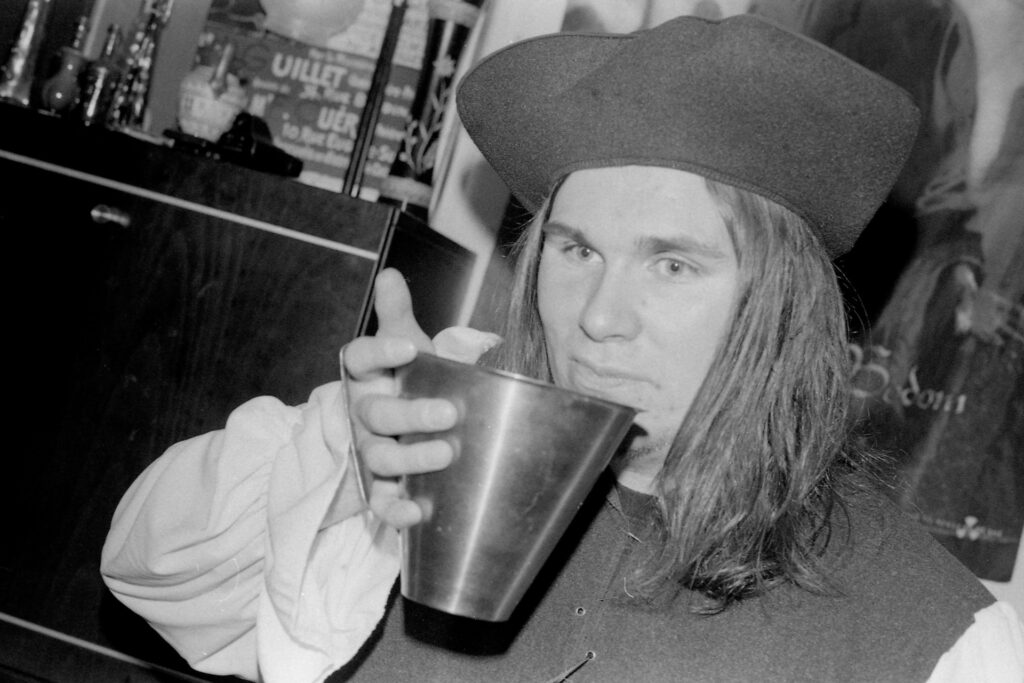 A black and white photograph of a man in his 20s wearing a pirate outfit. He's having a drink out of a large metal measurement cup.