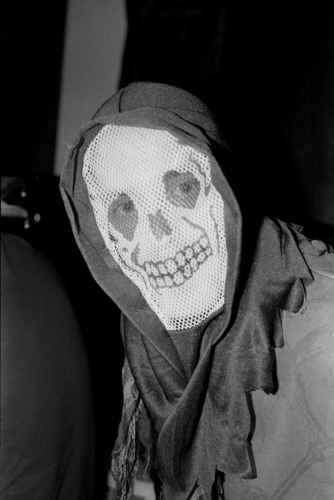 A black and white photograph of a person in Halloween costume. He's wearing a black hood and a mesh mask depicting a skull.