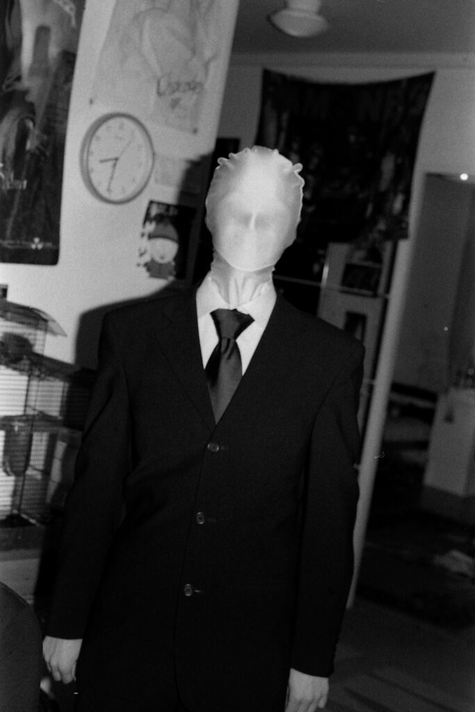 A black and white photograph of a person in a Halloween costume. They're wearing a black suit and a tie. Their head is covered with a featureless, morph-suit kind of mask.