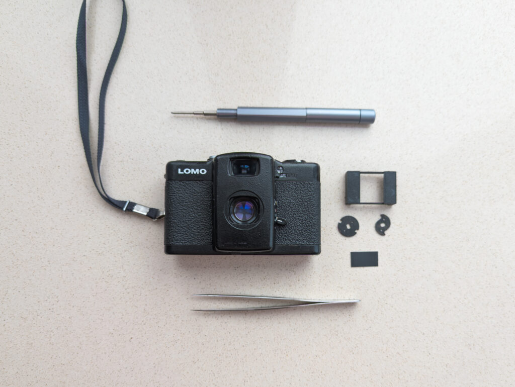 An overview photo of what's needed to create the new half-frame camera. We'll need: a camera, new parts, a screwdriver, tweezers, and a small piece of electrical tape.