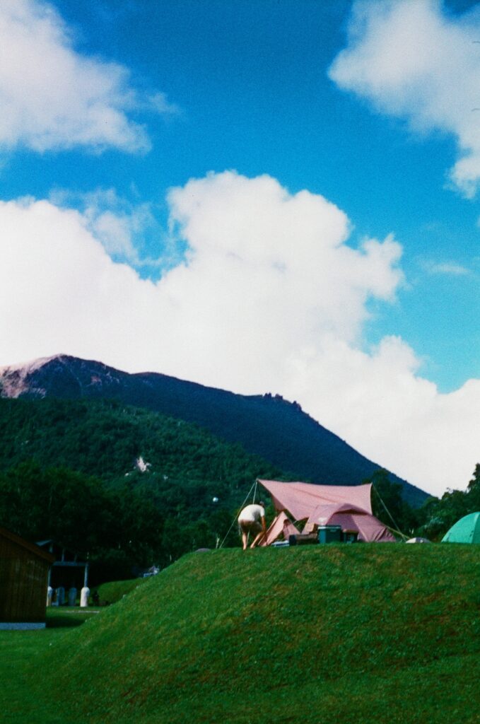 A man is setting up their tent on a hill. There are bright white clouds and green mountains in the background.